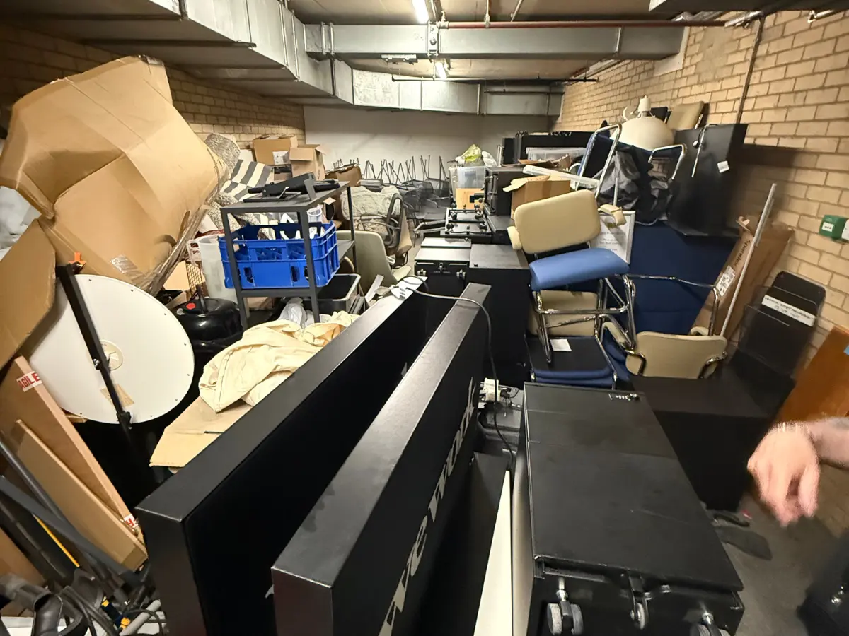 Crowded office storage area containing furniture, equipment and boxed items that need to be removed.